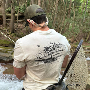 Angler streamside wearing the Angler's Coffee Mountain Scene tee with a landing net