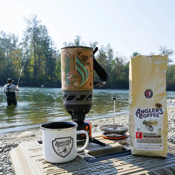 Caddis Blend coffee, Angler's enamel camp mug and camp stove on a gravel bar while an angler wades the river