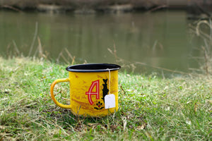 Angler's Coffee sachet brewing in the yellow Angler's camp mug by the river, tag over the rim
